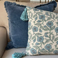 Two decorative throw pillows on a light beige sofa. The front pillow has a cream background with a blue floral vine pattern and a matching blue tassel, while the back pillow is a solid medium blue velvet with a ruffled edge and a matching tassel.