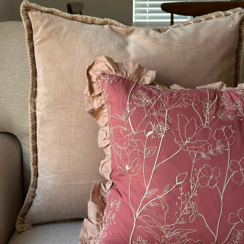 Two decorative throw pillows on a sofa: a large beige pillow with a ruffled edge in the back, and a dusty rose pillow with a white floral line pattern and ruffled trim in front.
