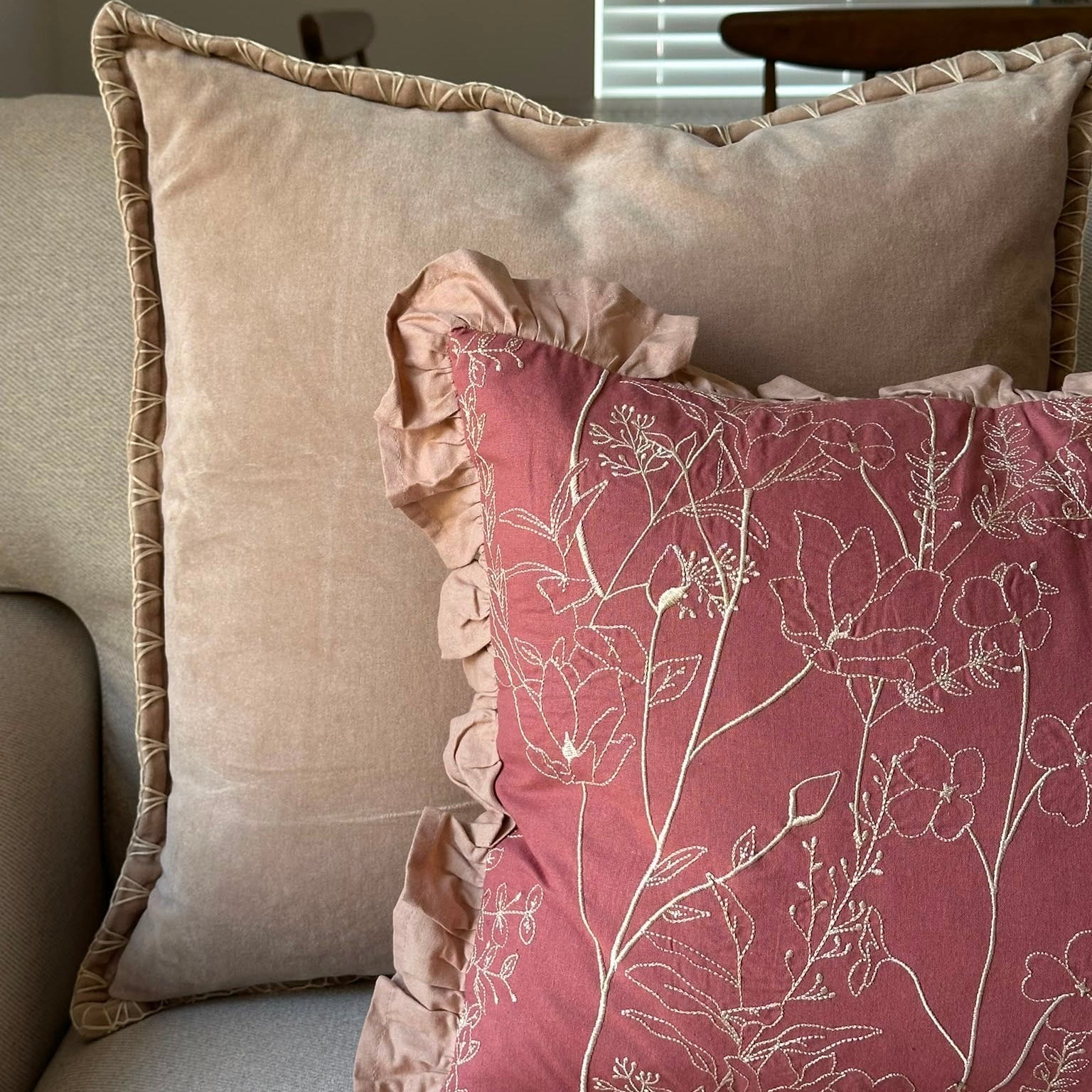 Two decorative throw pillows on a sofa: a large beige pillow with a ruffled edge in the back, and a dusty rose pillow with a white floral line pattern and ruffled trim in front.