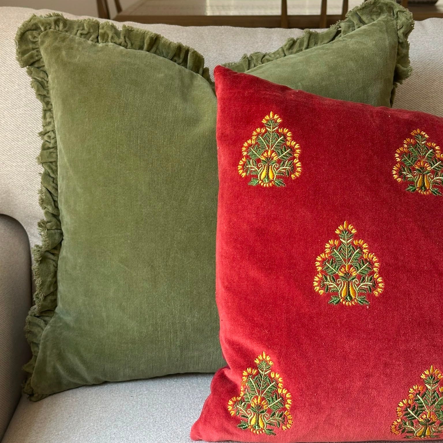 Two decorative throw pillows on a light gray sofa in a home setting. One pillow is sage green velvet with a ruffled edge, and the other is red velvet with embroidered gold and green floral motifs.