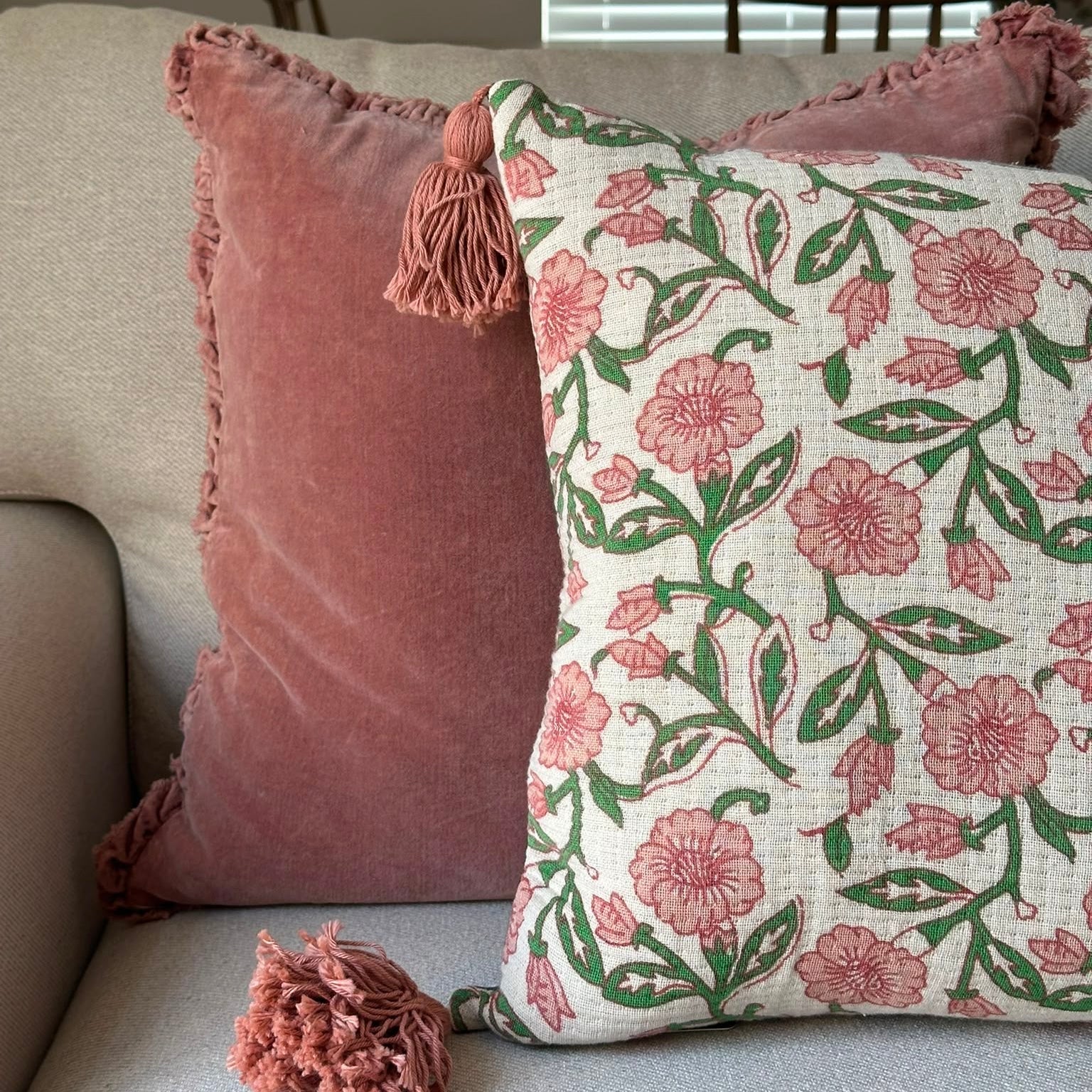 Two decorative throw pillows on a light gray sofa in a living room setting. One pillow is dusty rose pink velvet with a tassel, and the other is cream with a pink floral and green leaf pattern, with a matching tassel resting in front.