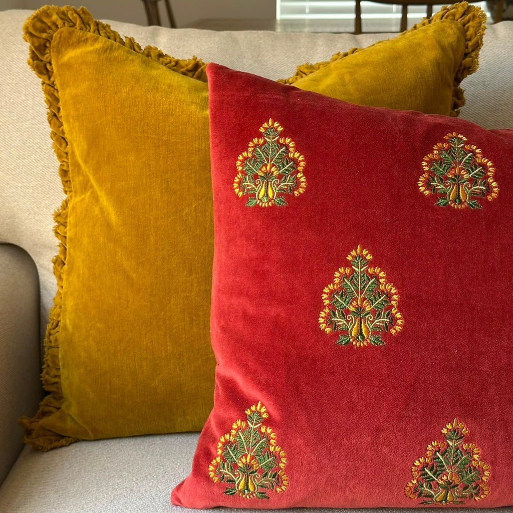 Two decorative throw pillows on a light-colored sofa: a mustard yellow pillow with fringe trim and a red pillow featuring green and gold embroidered motifs.