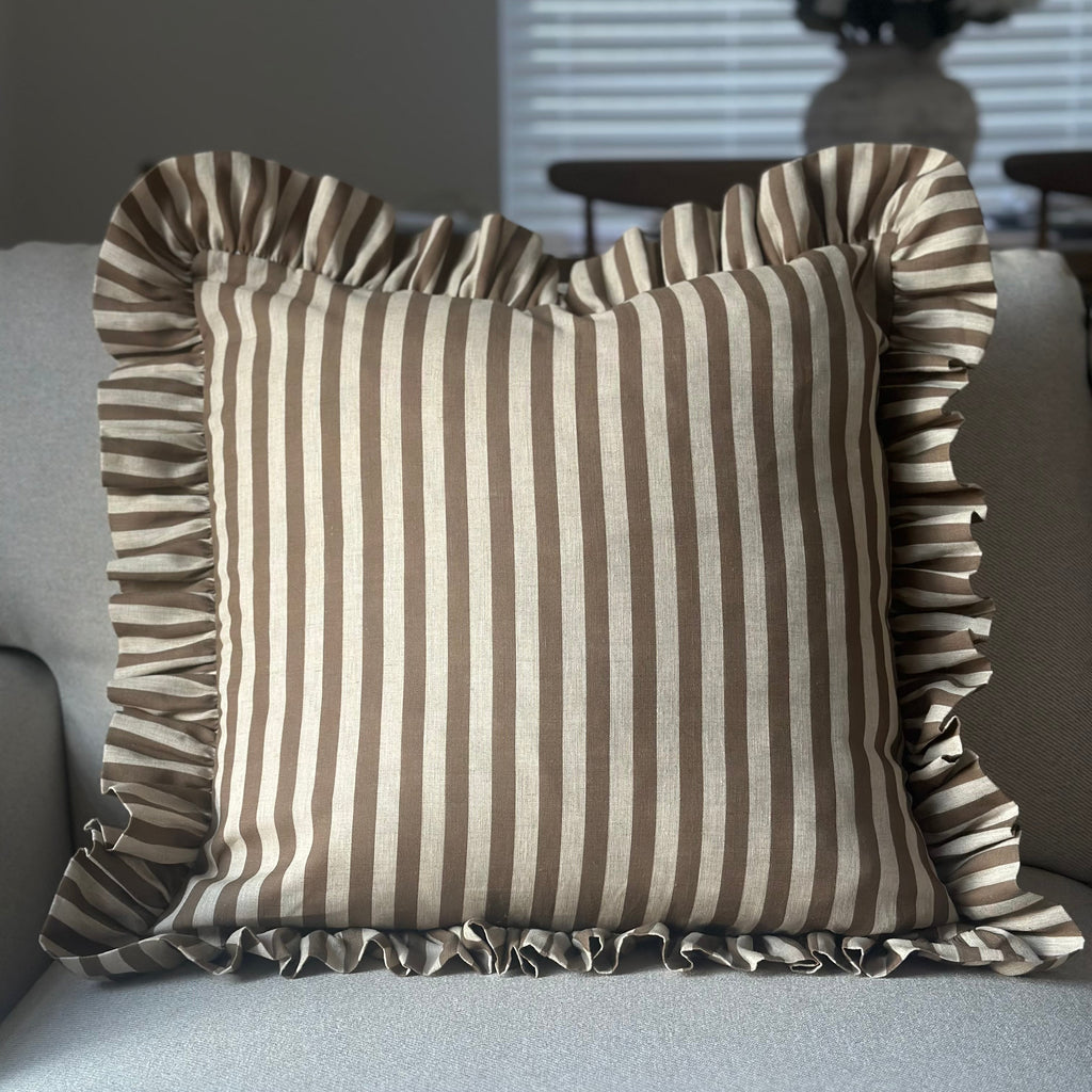A square throw pillow with vertical brown and beige stripes and ruffled trim, resting on a light gray sofa in a softly lit living room.