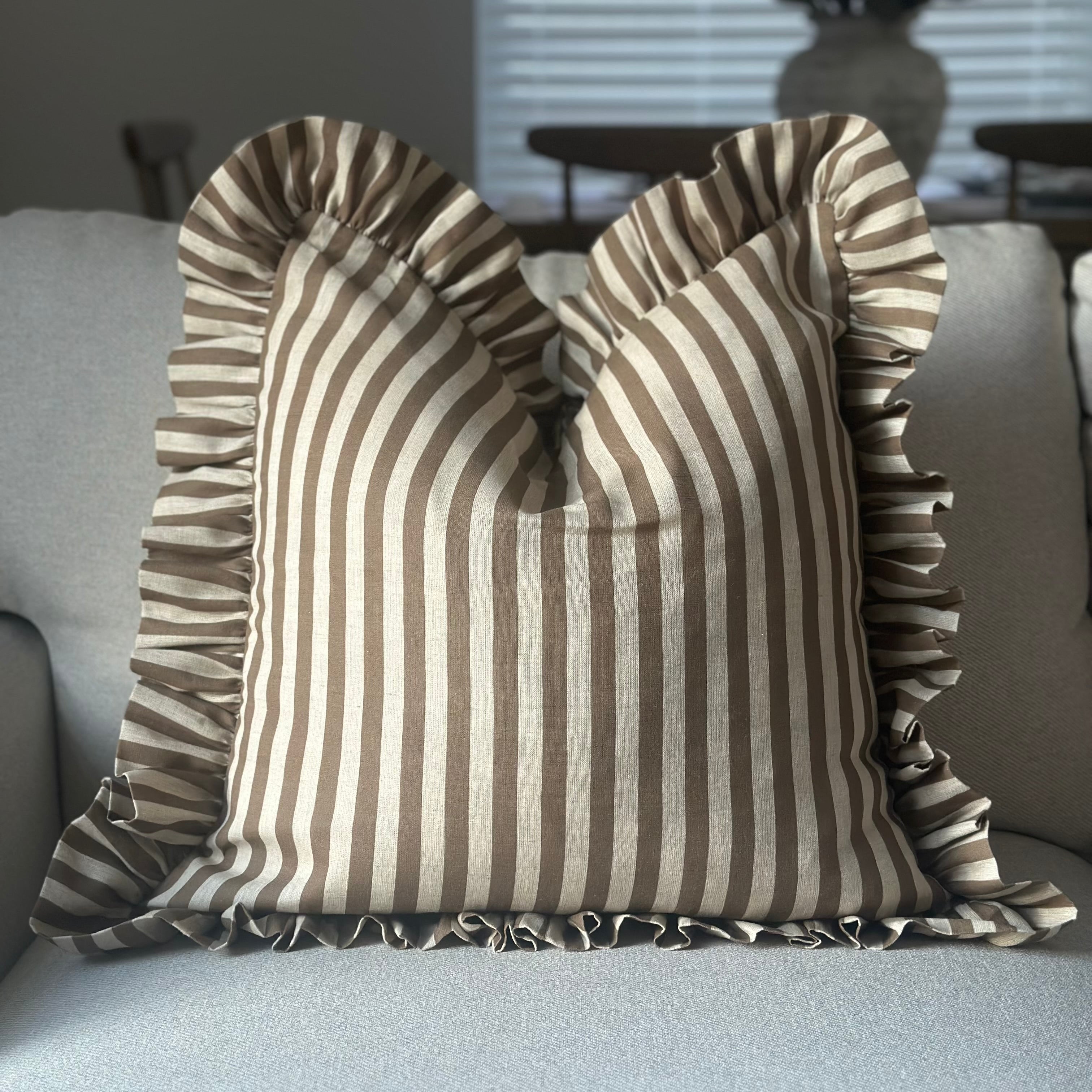 A square throw pillow with vertical brown and beige stripes and ruffled trim, resting on a light gray sofa in a living room setting.