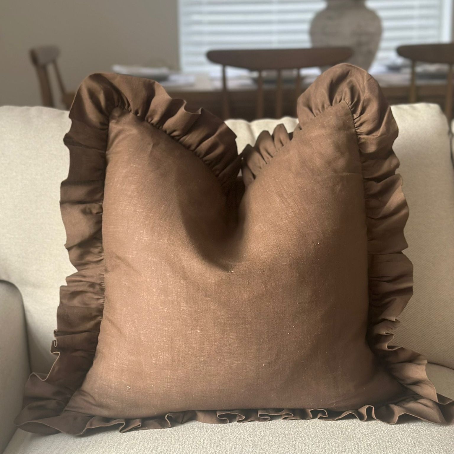 A square brown throw pillow with ruffled trim, resting on a light beige sofa in a softly lit living room.