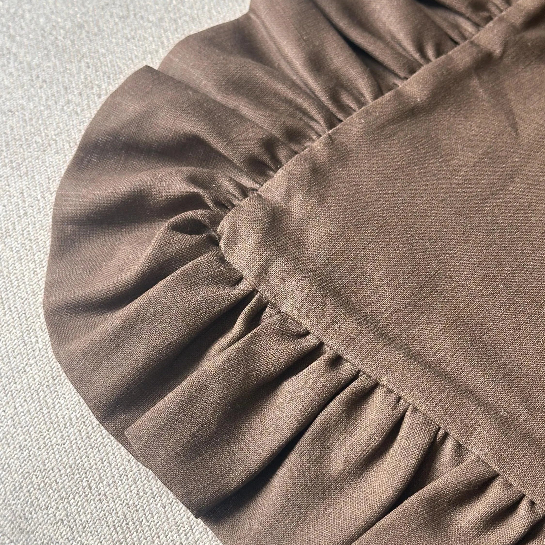 A close-up of a brown fabric product with a gathered ruffled edge, laid on a light gray textured surface.