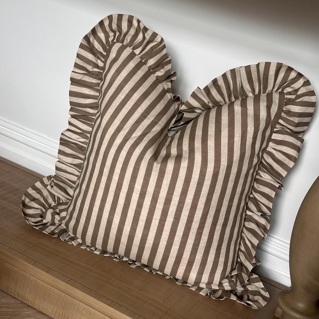 A square throw pillow with vertical brown and beige stripes and ruffled edges, resting on a wooden bench against a white wall.