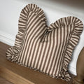 A square throw pillow with vertical brown and beige stripes and ruffled edges, resting on a wooden bench against a white wall.