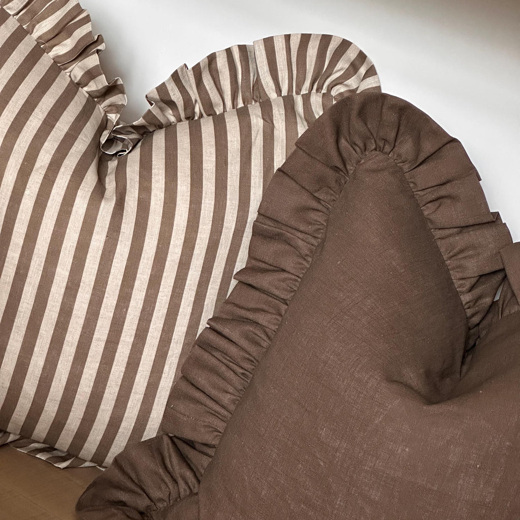 Close-up of decorative throw pillows with ruffled edges, including one in solid brown and one in brown and cream vertical stripes, resting on a light surface.